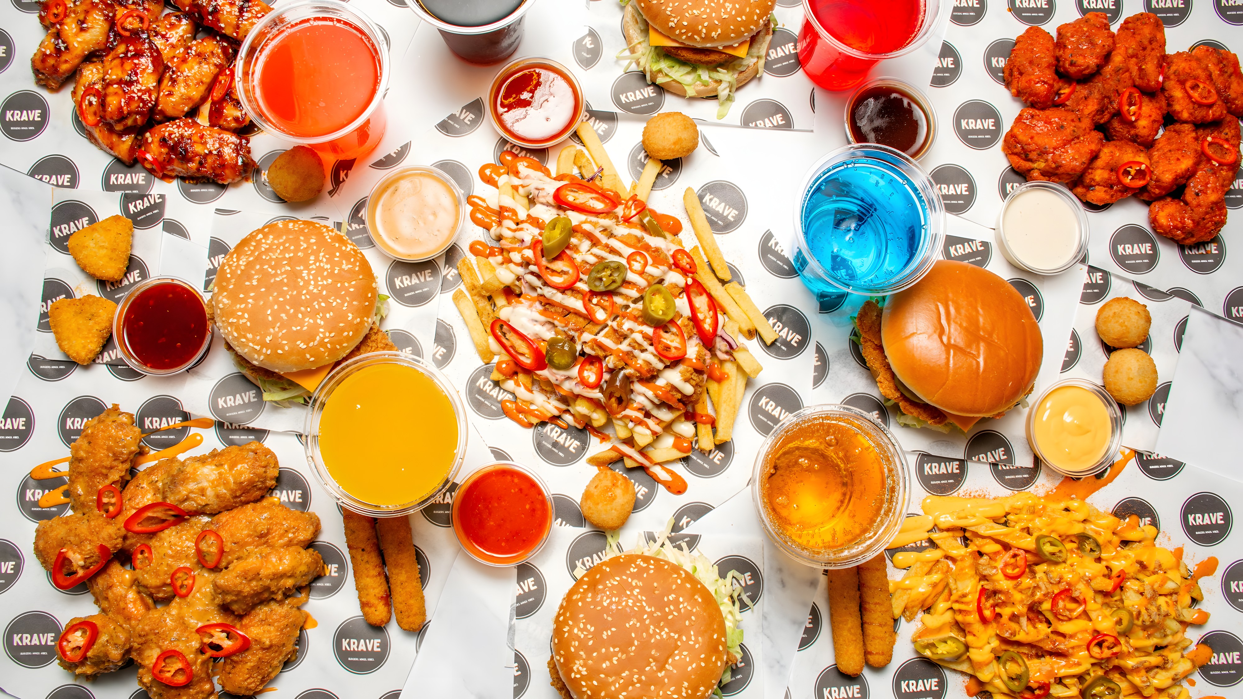 A full spread of KRAVE burgers, saucy wings, loaded fries and cold drinks laid out on branded paper